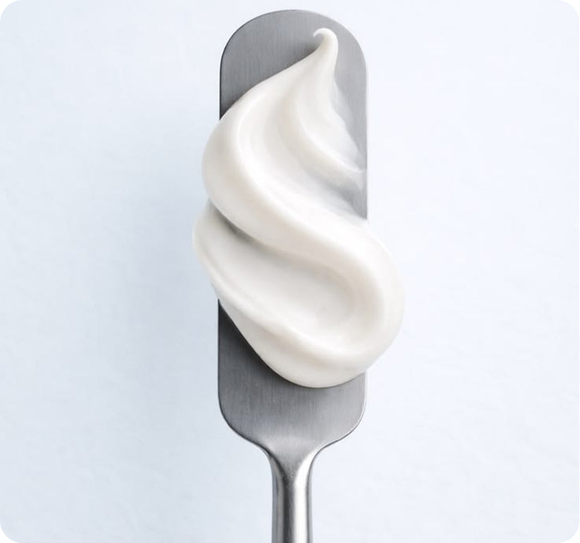 A thick, creamy white lotion or cream is swirled in an S-shape on a metallic silver spatula against a clean, light gray background.